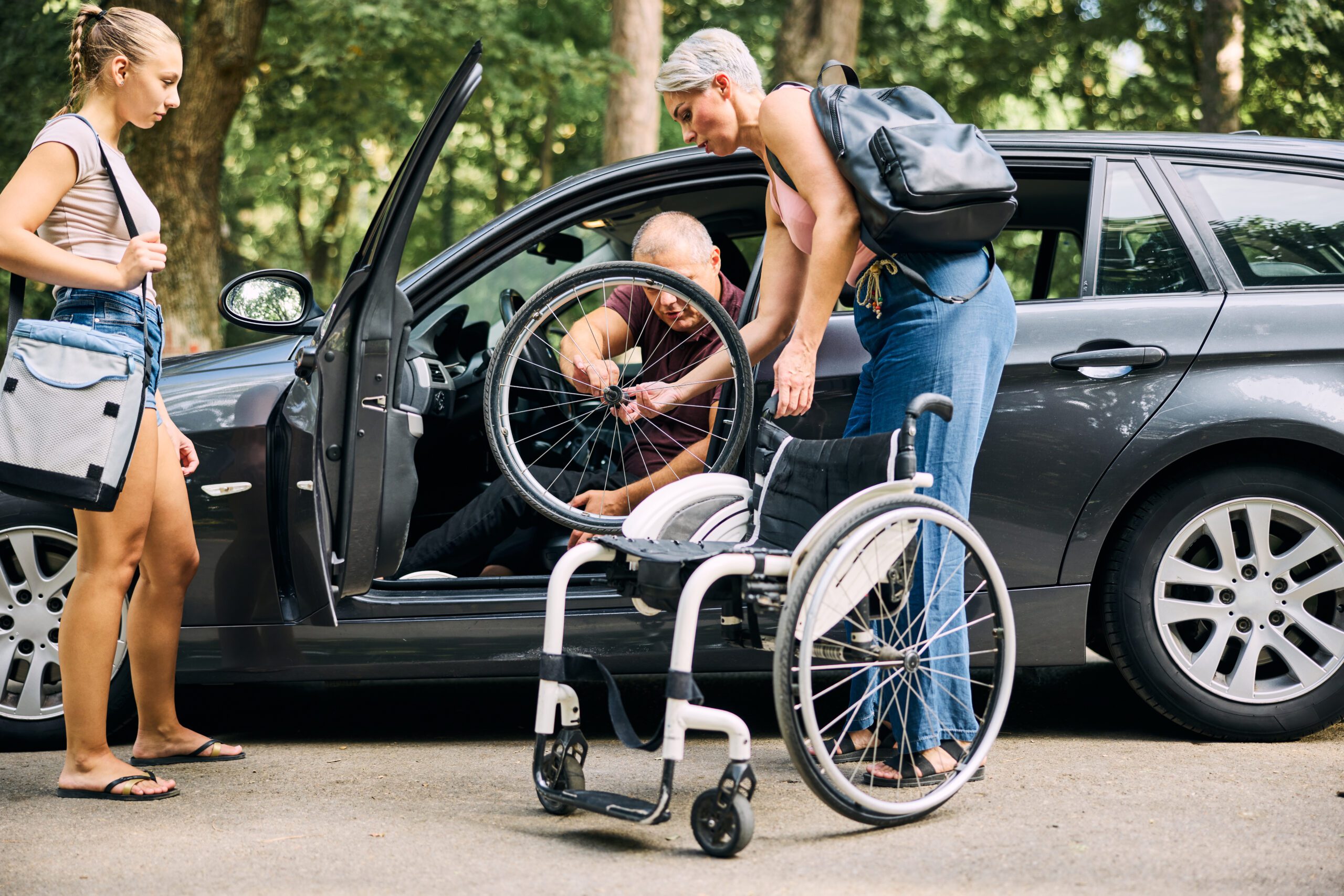 A family assists a man with a wheelchair into a car in a peaceful park setting.
