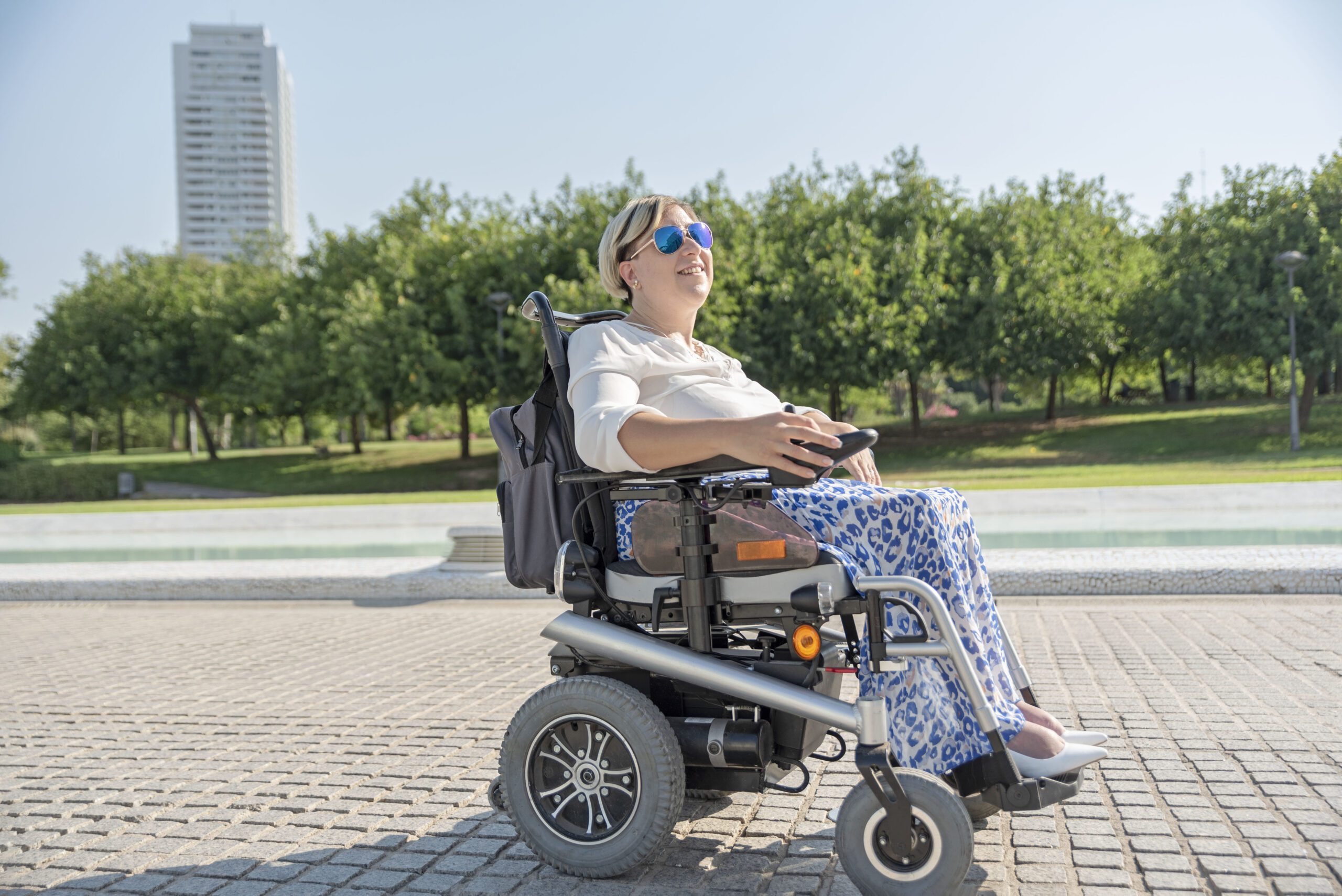 a smiling woman in an electric wheelchair with disability takes a stroll on a sunny day through an urban park in the city a smiling woman in an electric wheelchair with disability takes a stroll on a sunny day through an urban park in the city