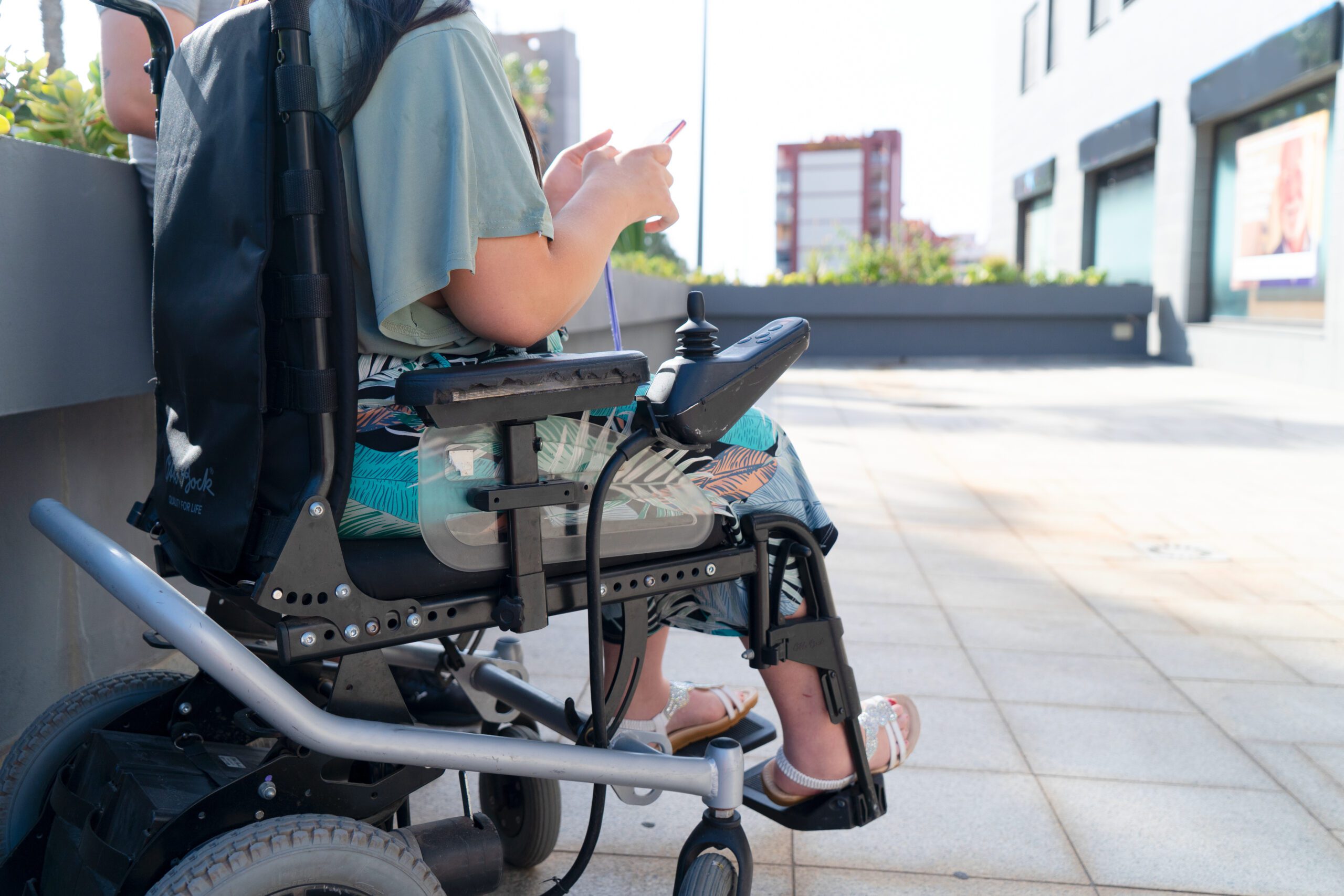 disabled handicapped woman in wheelchair using smartphone and smiling at camera while walking outdoors on sunny summer day. Disable people and modern technologies concept