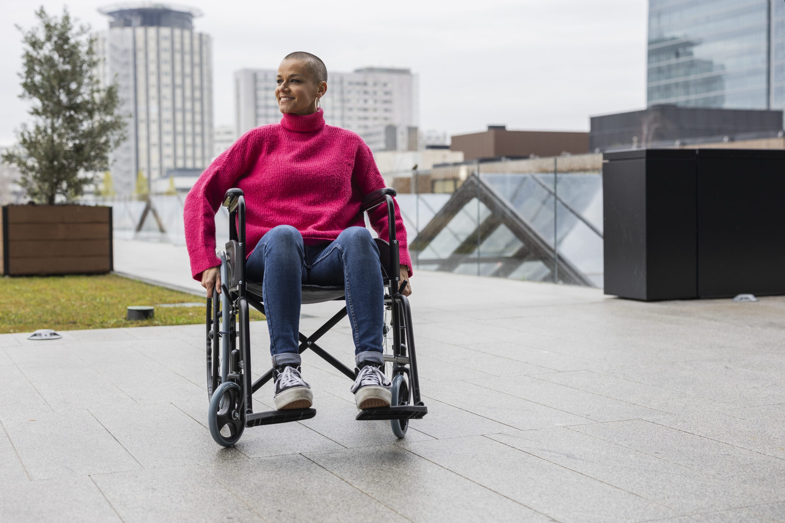 Happy young woman with a shaved head, dressed in a pink sweater and jeans, smiling while moving in a wheelchair along a vibrant city street, embracing her independence and positivity